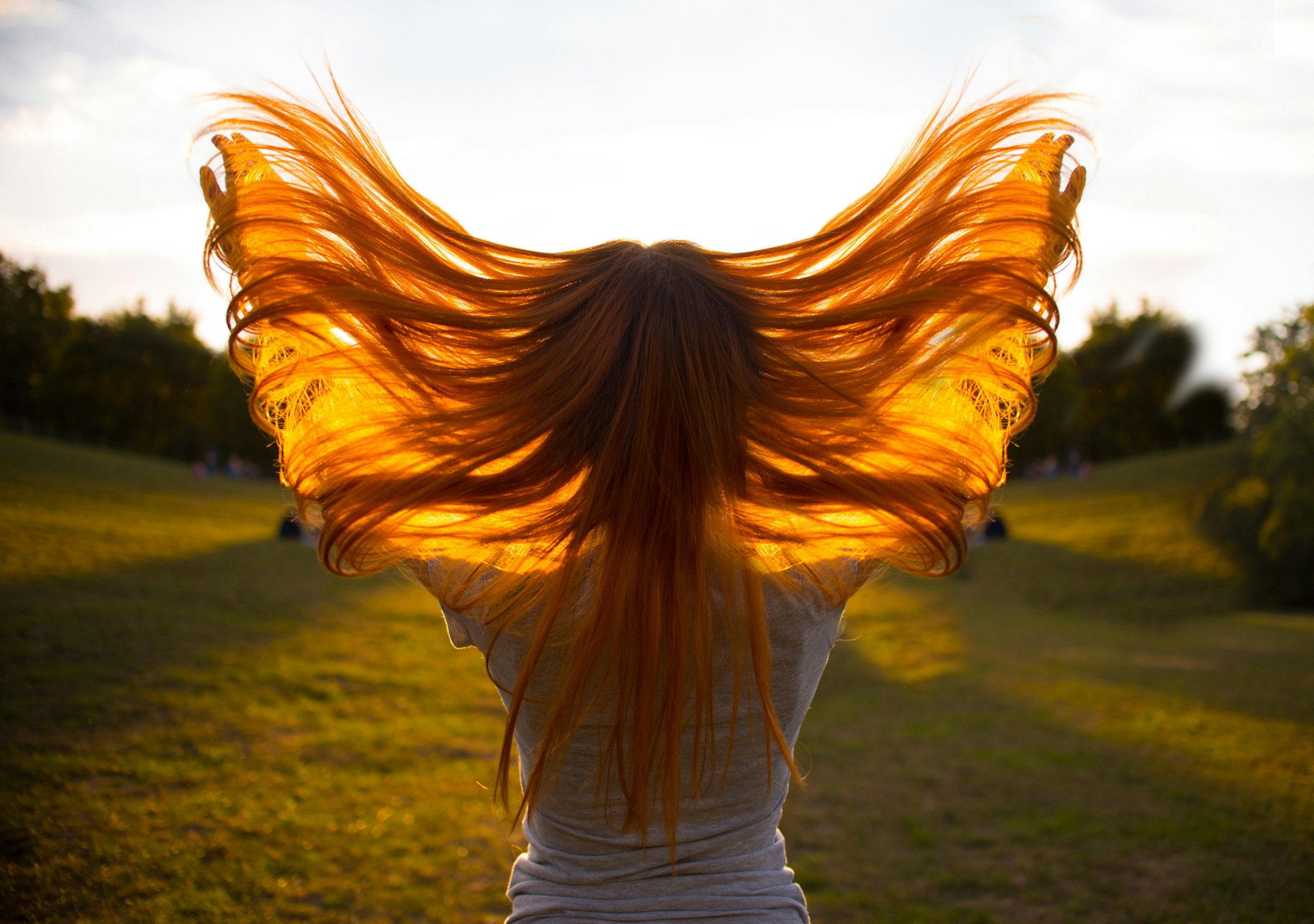 Femme de dos faisant voler ses longs cheveux cuivrés, illuminés par un contre-jour doré en extérieur