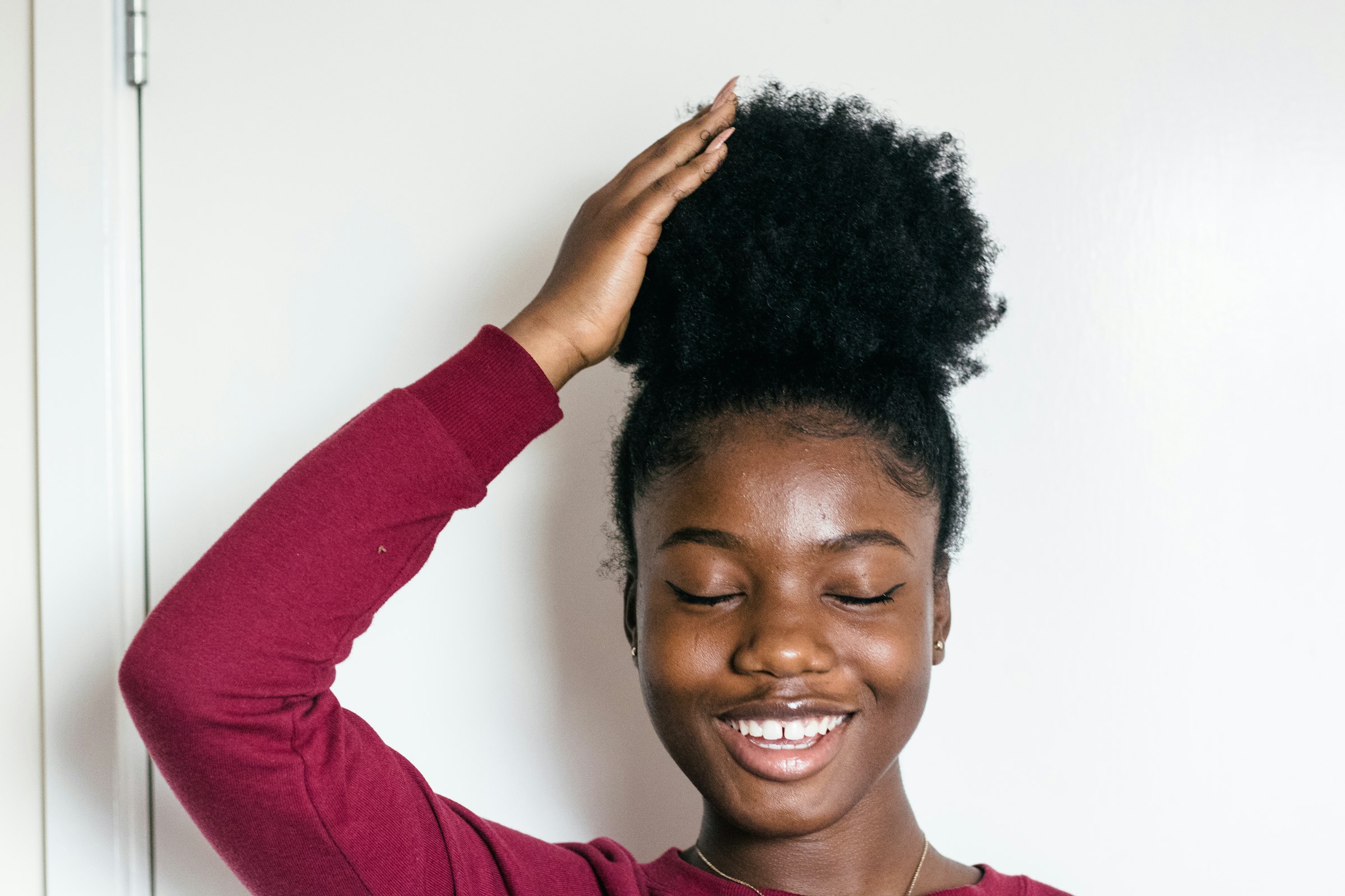 Jeune femme noire aux cheveux crépus naturels relevés en chignon afro, la main posée sur ses cheveux, souriante devant un mur blanc lumineux