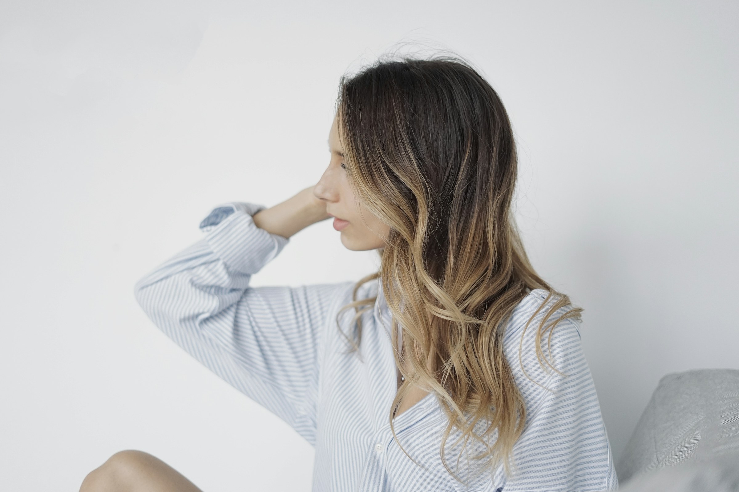 Femme de profil aux cheveux ondulés châtain aux pointes éclaircies, chemise claire sur fond blanc lumineux