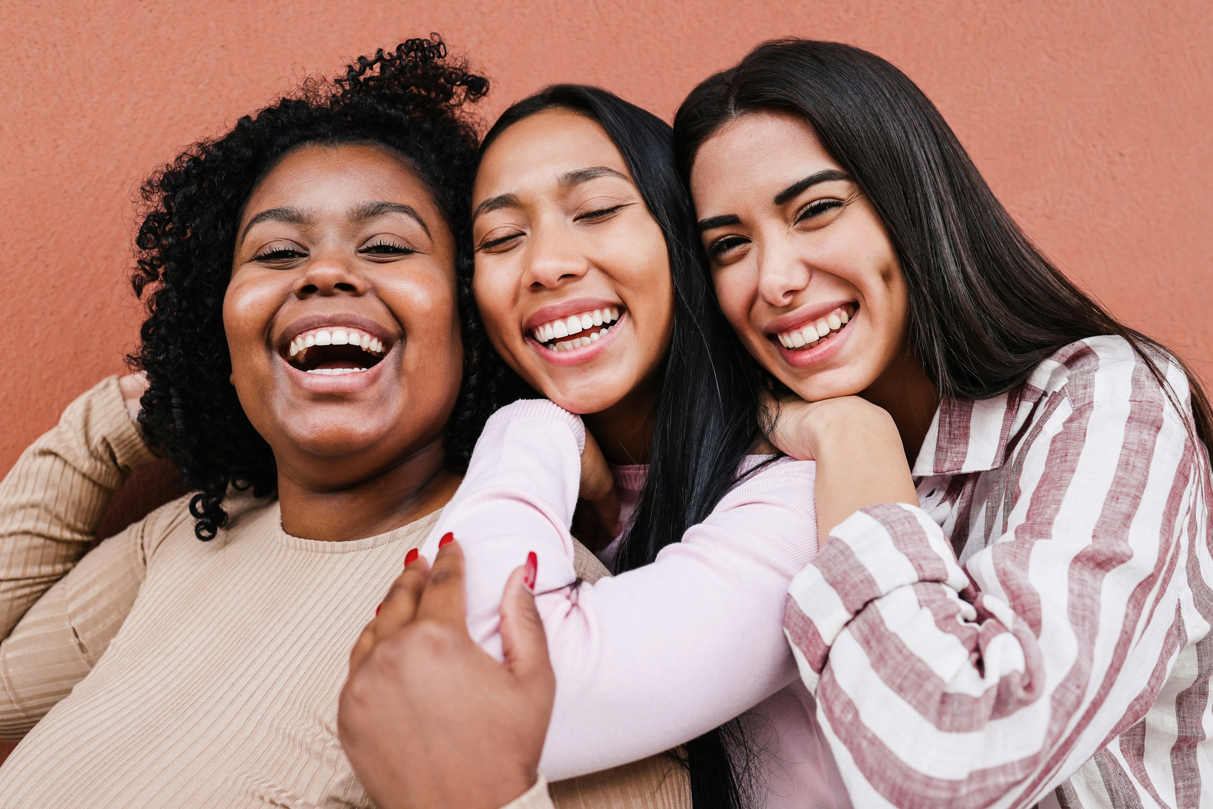 Trois femmes souriantes d'origines différentes, cheveux bouclés et cheveux longs lisses, réunies sur un fond terracotta chaleureux